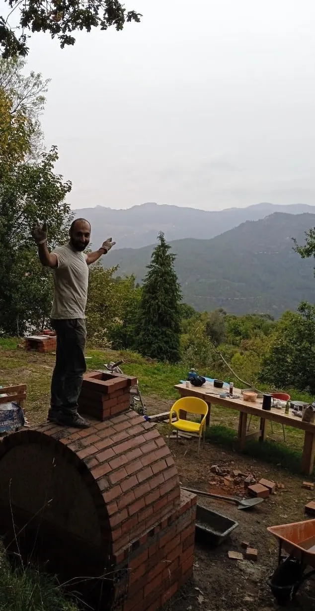 Shahin standing on top of the brick pizza oven he is building, mountains behind
