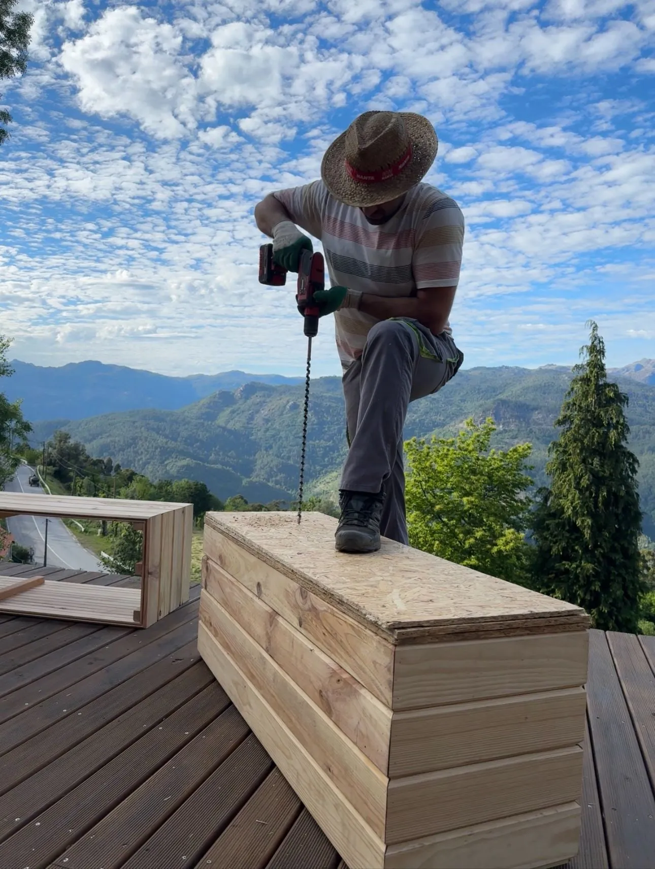 Shahin in a straw hat drilling a wooden storage bench on a deck